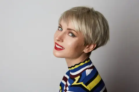 Kobieta z rudymi piegami i krótką, blond pixie bob fryzurą, uśmiecha się, nosząc kolorowy sweter.