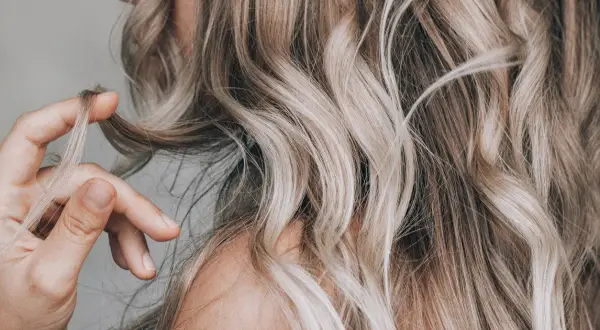 Dłoń dotyka falujących włosów w odcieniach ombre blond, tworząc subtelny efekt.